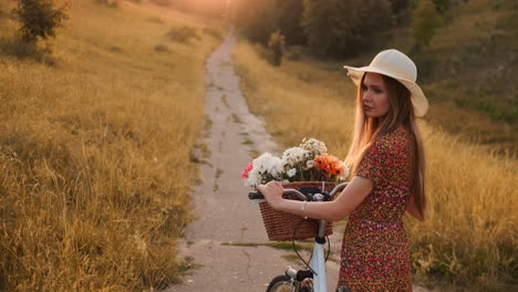 Back-kind-of-Happy-blonde-girl-in-dress-and-hat-turns-around-and-smiling-cheerfully-looks-at-the-camera-and-flirts-strolling-around-the-field-in-summer-with-bike-and-flowers.